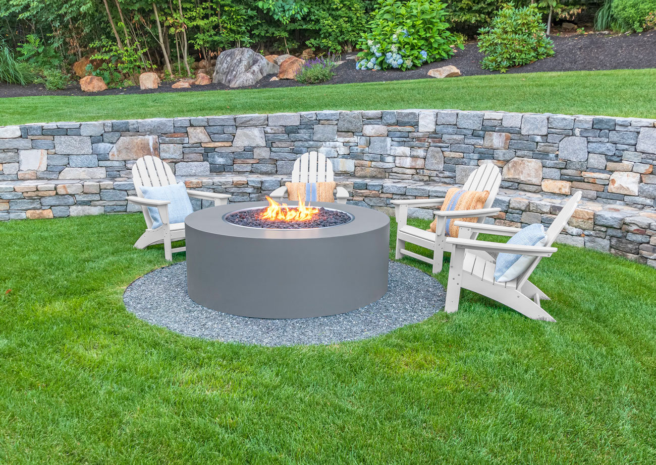 Natural stone retaining wall holding a planted grade above a lawn, with a fire feature and seating on the level below