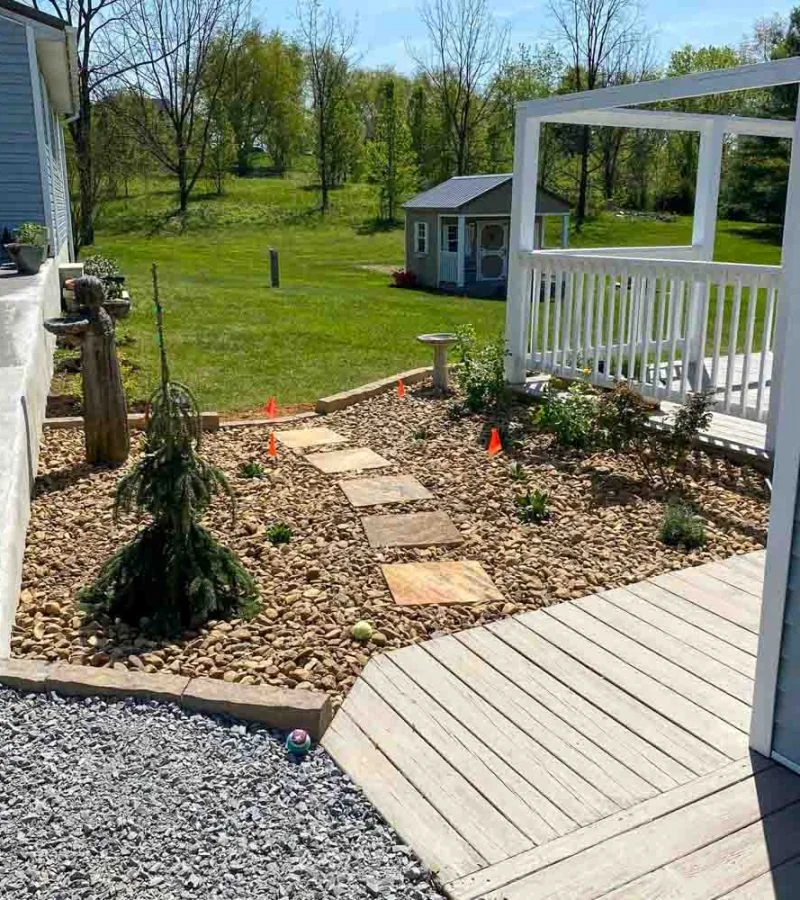 Decorative landscape bed featuring river rock ground cover, flagstone stepping stones, newly planted shrubs and small evergreen tree, wooden deck with white pergola, birdbath, and orange placement markers